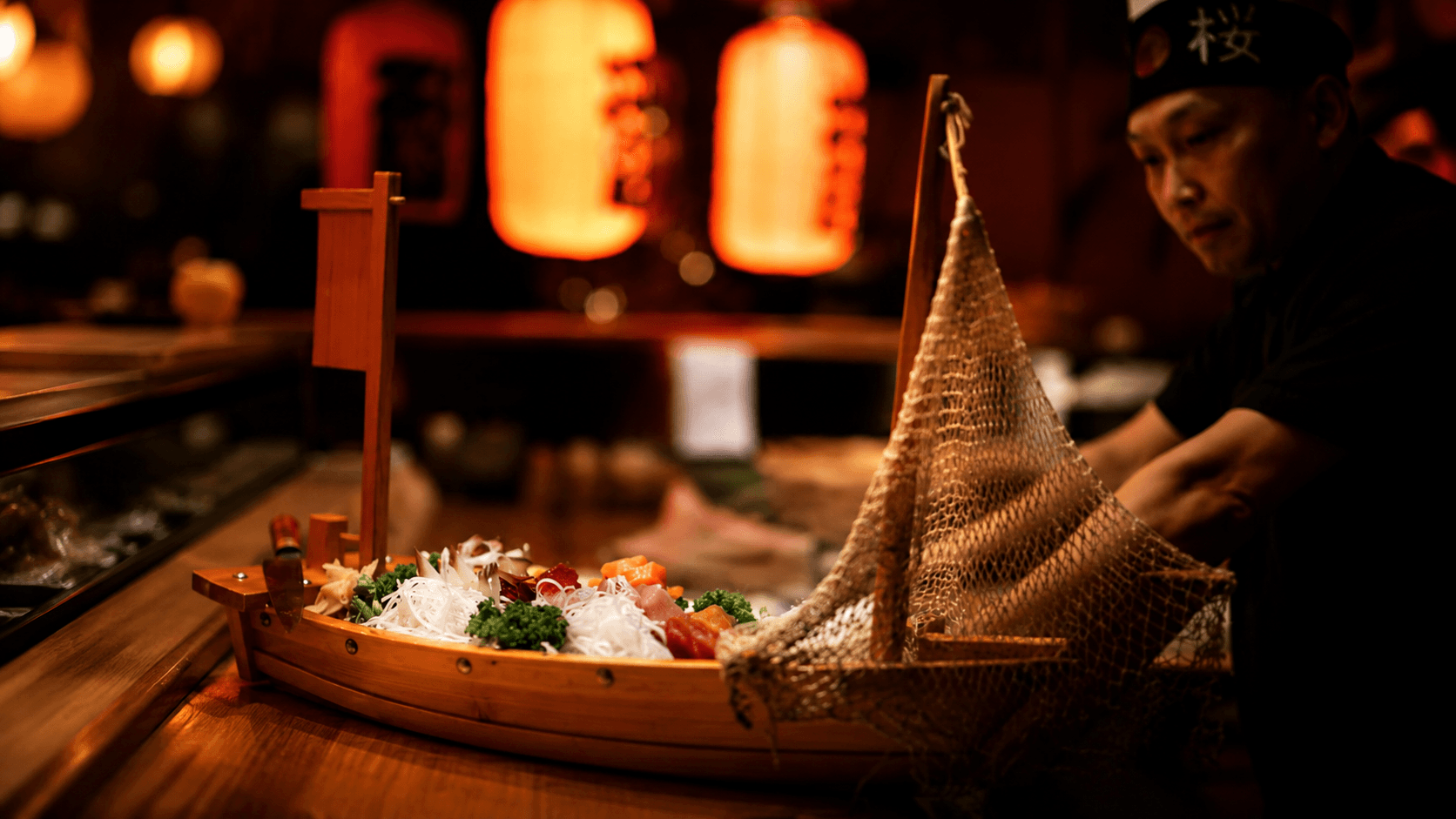 A chef plating a wooden sushi boat at Tenkuu, beneath warm red lanterns in a dark izakaya